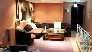 Ein Wohnzimmer mit einer Couch, einem Couchtisch und einem Fernseher. Ein Fenster mit Vorhängen, ein Heizkörper und eine Tür sind ebenfalls vorhanden.