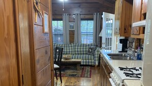 Interior acolhedor de uma cabana com sofá estampado em xadrez, piso de madeira, cozinha com fogão e ventilador de teto.