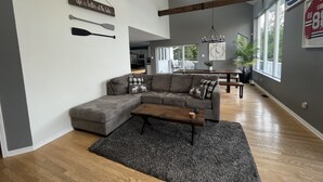 Ein geräumiges Wohnzimmer mit einem großen, grauen Ecksofa, einem Holztisch und einem grauen Teppich. Es gibt eine Essecke mit einer Holzbank und einer Pendelleuchte. Der Raum hat freiliegende Holzbalken, große Fenster und einen Blick ins Freie.