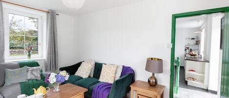 A living room with a green sofa, a coffee table, and a lamp. There is a window with curtains and a view of greenery outside.