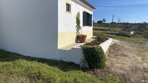Une maison de plain-pied à l’extérieur blanc et au rez-de-chaussée jaune, avec une petite fenêtre dotée de volets noirs et un pot de fleurs placé à côté de l’entrée.