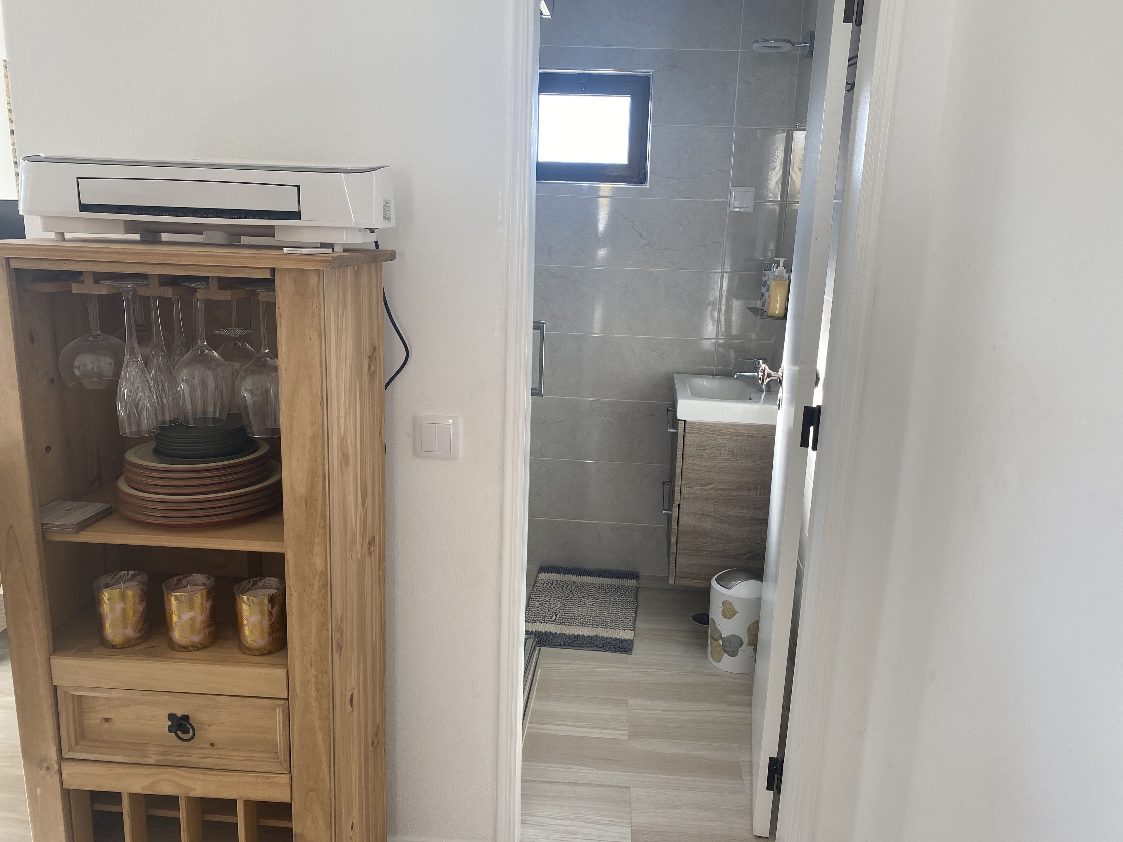 Une pièce avec un buffet en bois contenant des verres à vin et des assiettes, un mur blanc avec une fenêtre, et une salle de bain avec un lavabo et des murs carrelés.