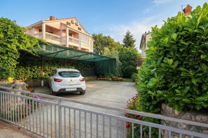 Un'auto bianca parcheggiata di fronte a un edificio con un balcone, circondato da vegetazione e fiori.