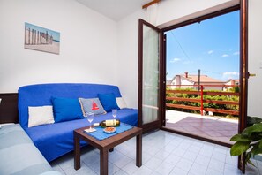 Quarto com um sofá azul, uma mesa pequena com uma garrafa e copos, e vista para um jardim e prédios através de uma porta de vidro.
