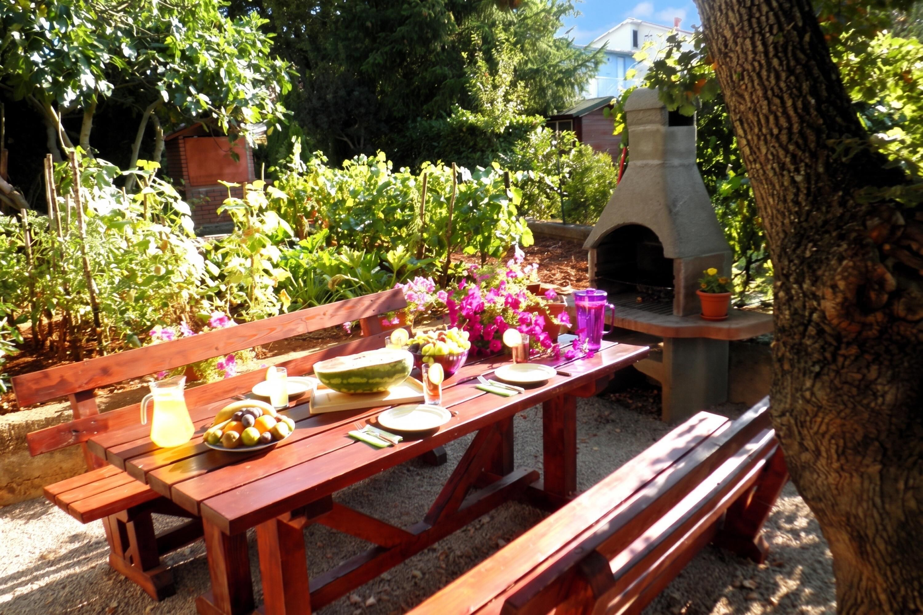 A wooden picnic table set outdoors with a pizza oven, surrounded by greenery and a building in the background.