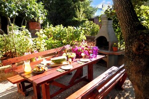 Conjunto de mesa e bancos de madeira para piquenique ao ar livre, com um forno a pizza, rodeado por vegetação e um edifício ao fundo.