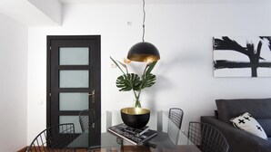 Un comedor moderno con una mesa de vidrio, sillas negras, una lámpara colgante negra y dorada, una planta en maceta y un sofá negro con un cojín blanco.