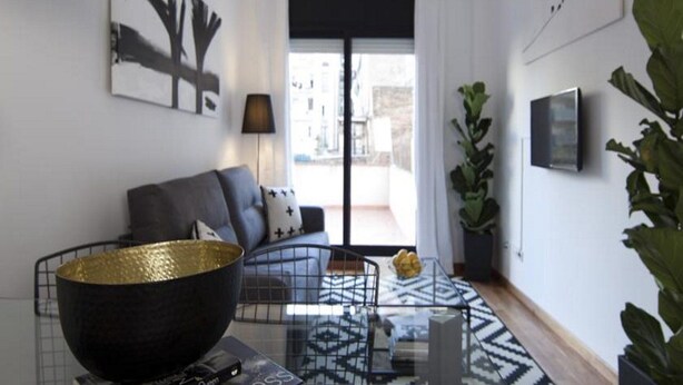 Una sala moderna con un sofá, un bol negro grande, una mesa de centro de vidrio y vista a la calle por una ventana amplia.