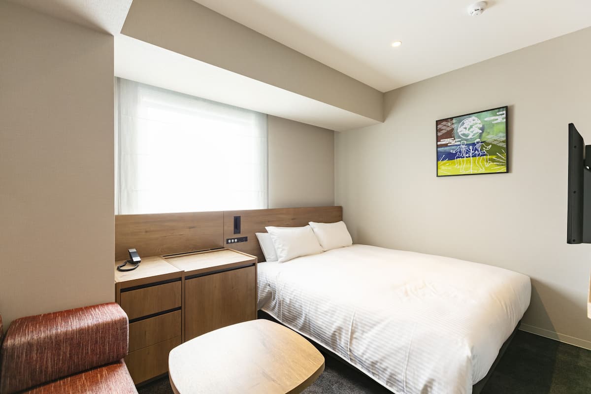 Una habitación de hotel moderna con una cama grande, un escritorio de madera, un sillón con cojín, una mesita y una ventana con persianas.