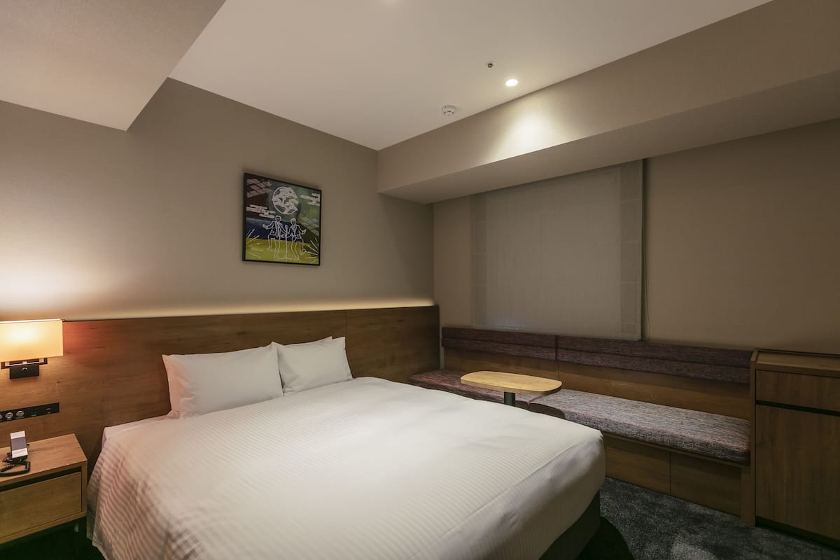 Una habitación de hotel con cama, mesita de noche, lámpara, banco y un cuadro en la pared.