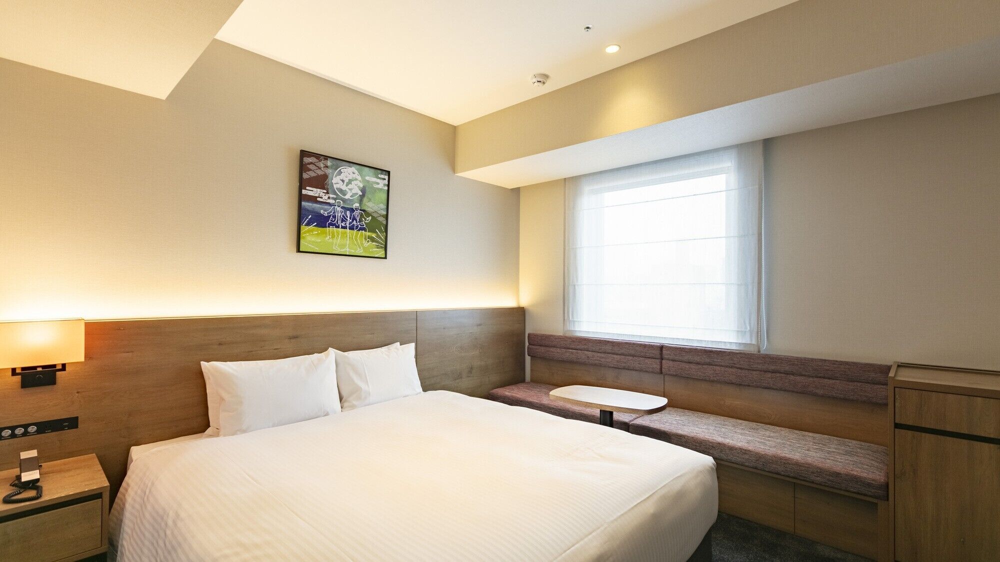 A hotel room with a large bed, bedside table, lamp, and a window with blinds.