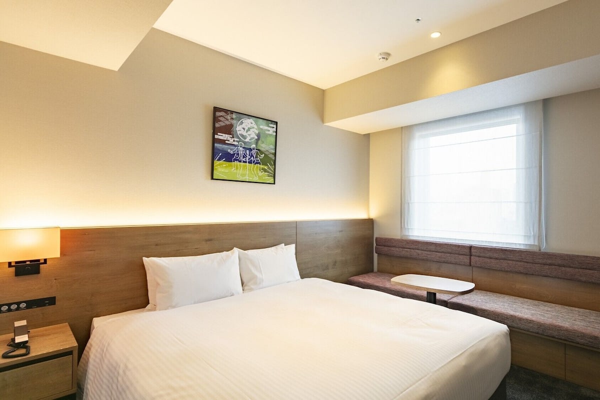 Habitación de hotel con una cama grande, mesita de noche, lámpara y ventana con persianas.