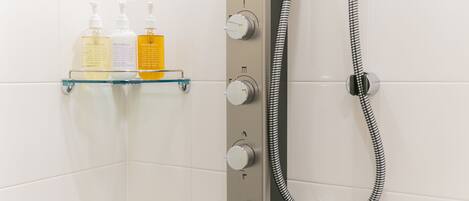 A shower with control knobs and a handheld showerhead, and a glass shelf with toiletries.