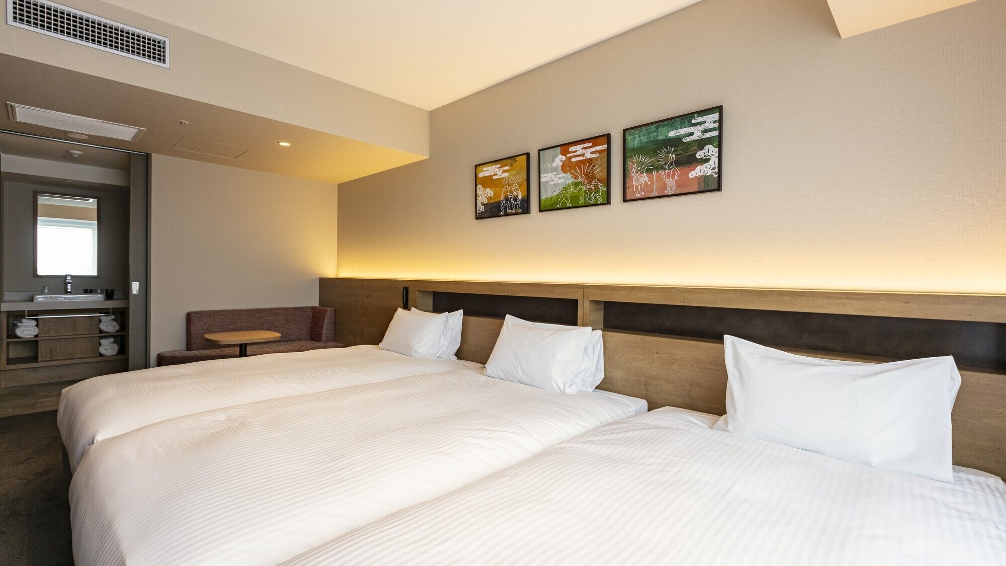 A hotel room with a large bed, a small table, a sofa, a bathroom with a sink, and three wall-mounted artworks.