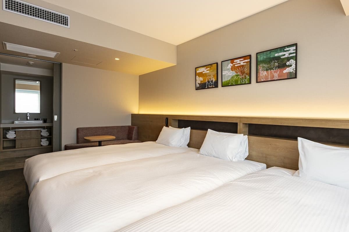 Habitación de hotel con una cama grande, una mesita pequeña, un sofá, un baño con lavamanos y tres obras de arte colgadas en la pared.
