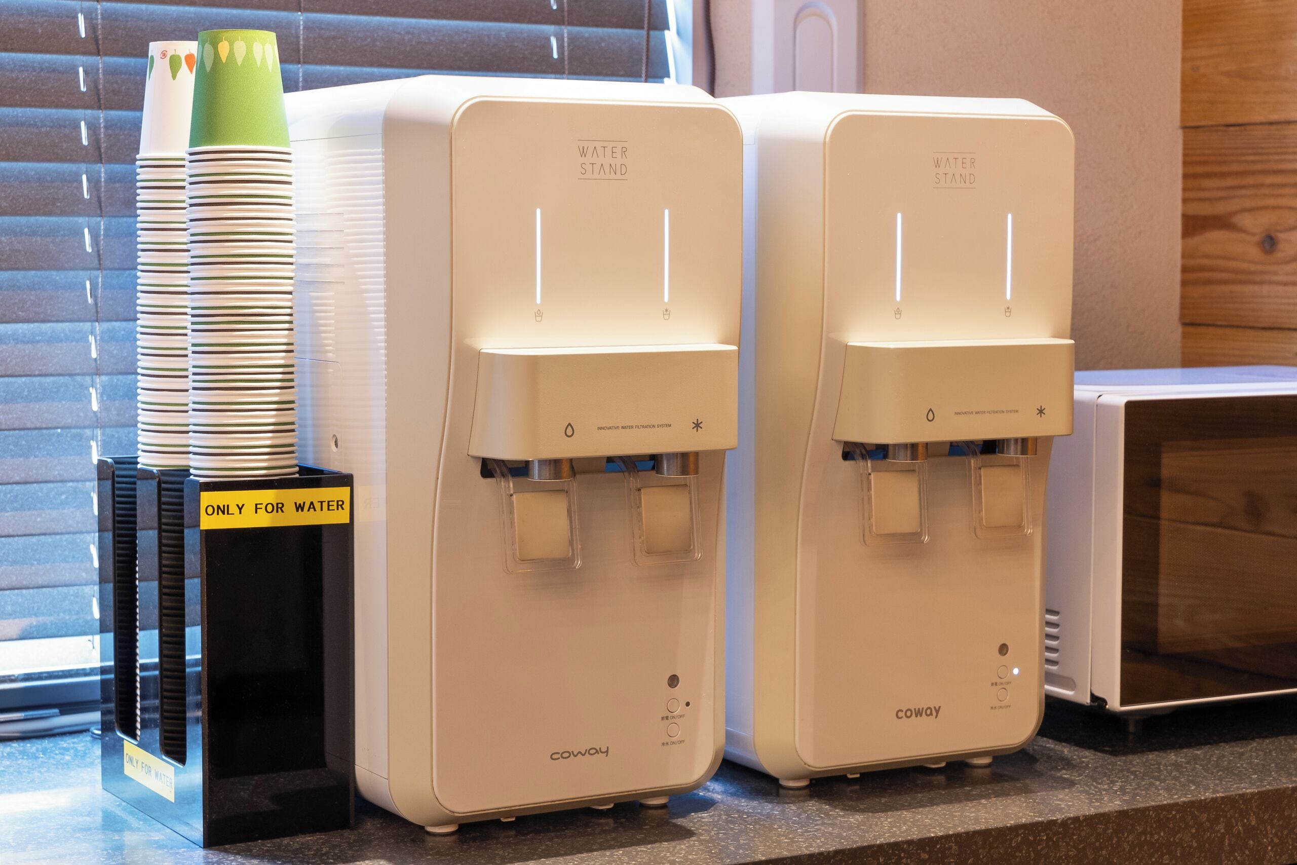 Dos dispensadores de agua con el rótulo 'WATER STAND', un cartel que indica 'SOLO AGUA', platos apilados y un sistema de filtración de agua Coway.