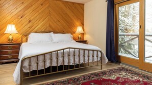 A bedroom with a bed, two bedside lamps, a wooden headboard, a red patterned rug, and a view of snow-covered trees through a glass door. - CPE1-Lovely Chalet! A/C, Pets Ok! (Davis)