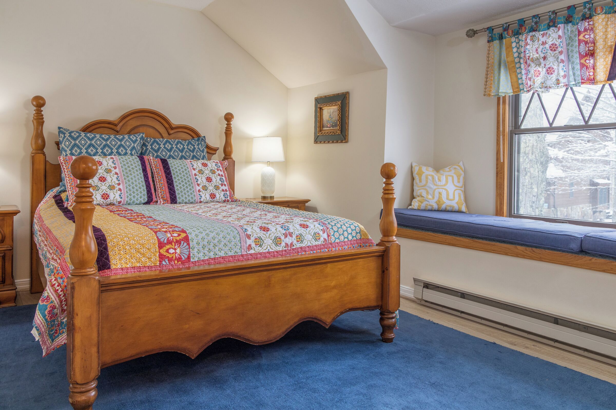 A bedroom with a wooden four-poster bed, a blue carpet, a window with a view, and a bedside table with a lamp.