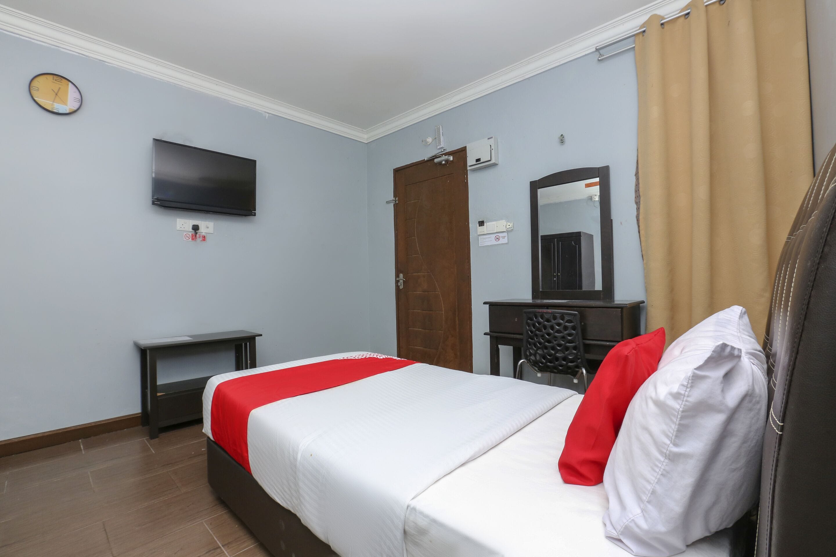 A hotel room with a bed, a desk, a chair, a TV mounted on the wall, a clock, and a door.