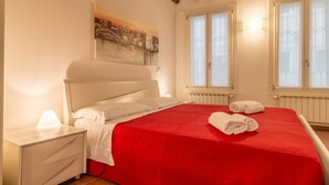 A bedroom with a red bedspread, white pillows, a nightstand with a lamp, and a painting on the wall.