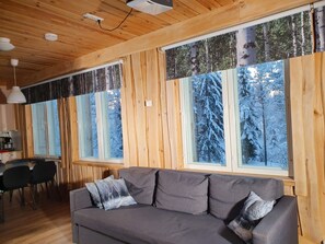 L'intérieur d'une cabane avec un plafond en bois, un canapé gris agrémenté de coussins et de grandes fenêtres donnant sur une forêt enneigée.