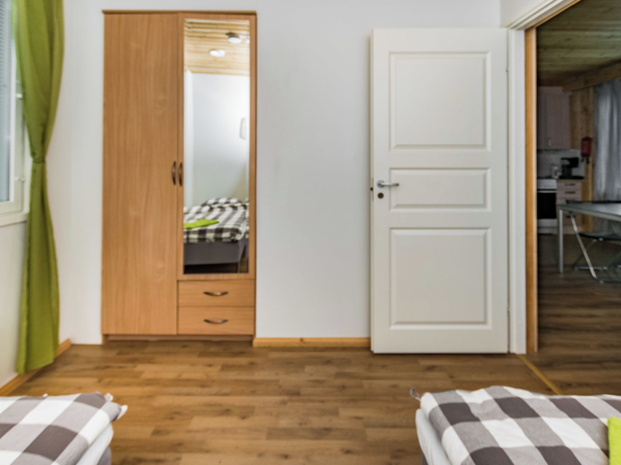 A room with a wooden wardrobe, a bed with checkered bedding, and a door leading to another room.