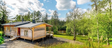 Une cabane en bois avec une véranda, une petite terrasse et un étang en arrière-plan.
