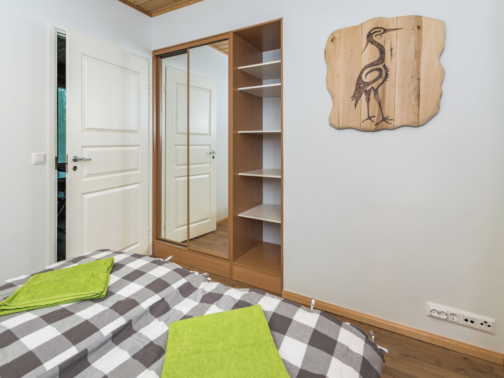 A bedroom with a checkered bedspread, a green towel, a wooden wardrobe, and a wall-mounted shelf with a bird design.