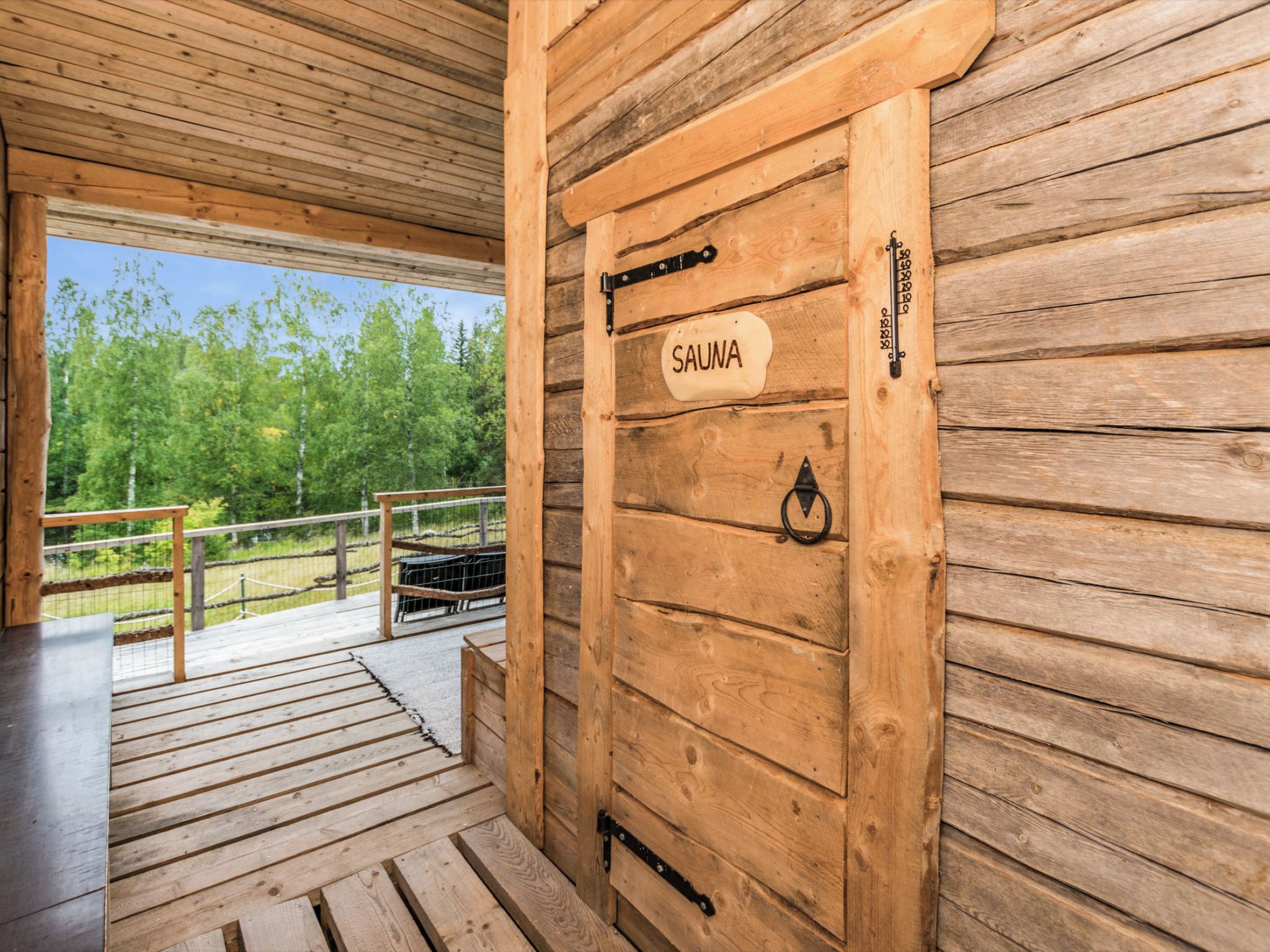 A wooden door labeled 'SAUNA' with a metal handle and a metal latch, leading to an outdoor deck with a railing.