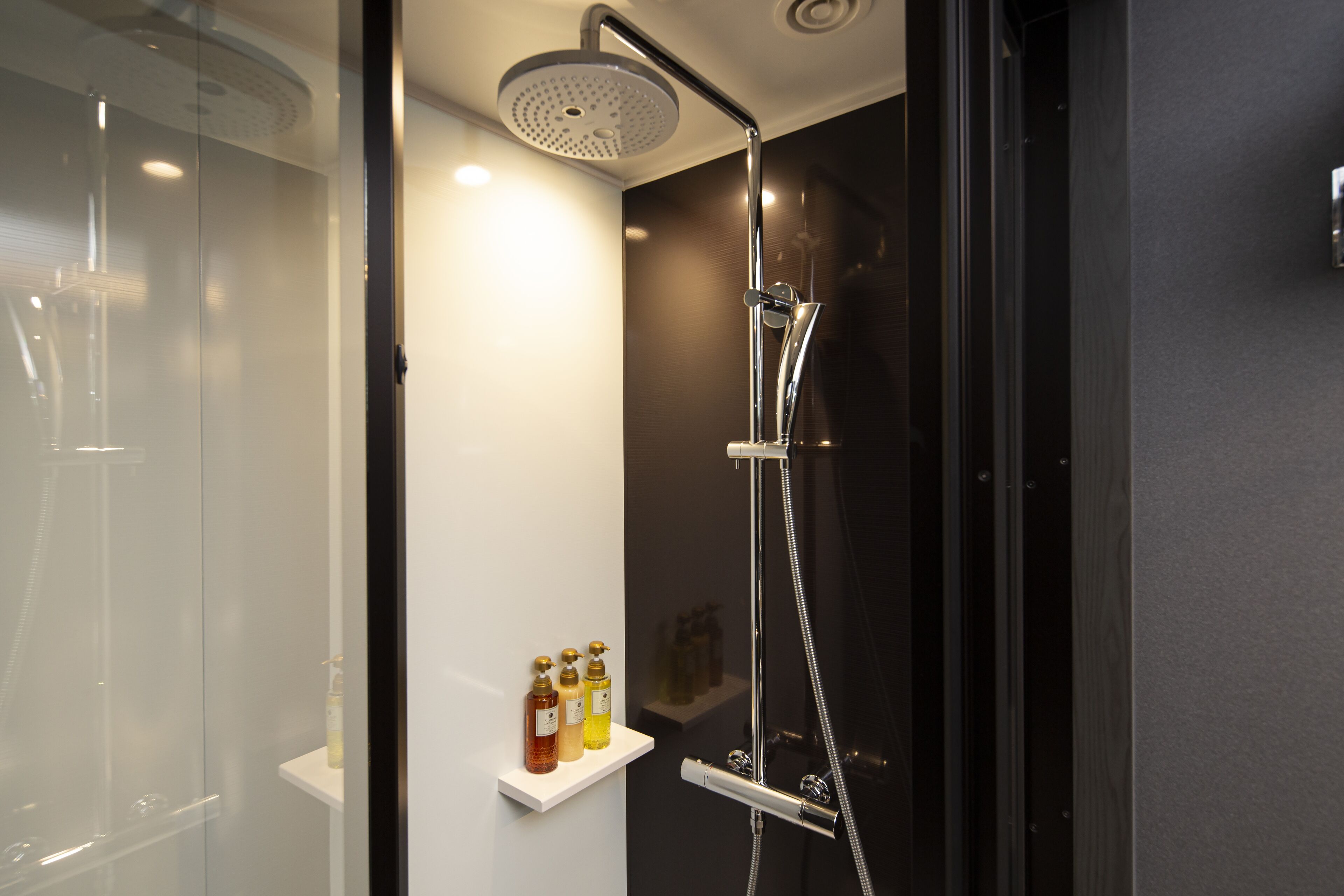 A modern shower with a handheld showerhead and a wall-mounted faucet. There are three bottles of shampoo on a small shelf.