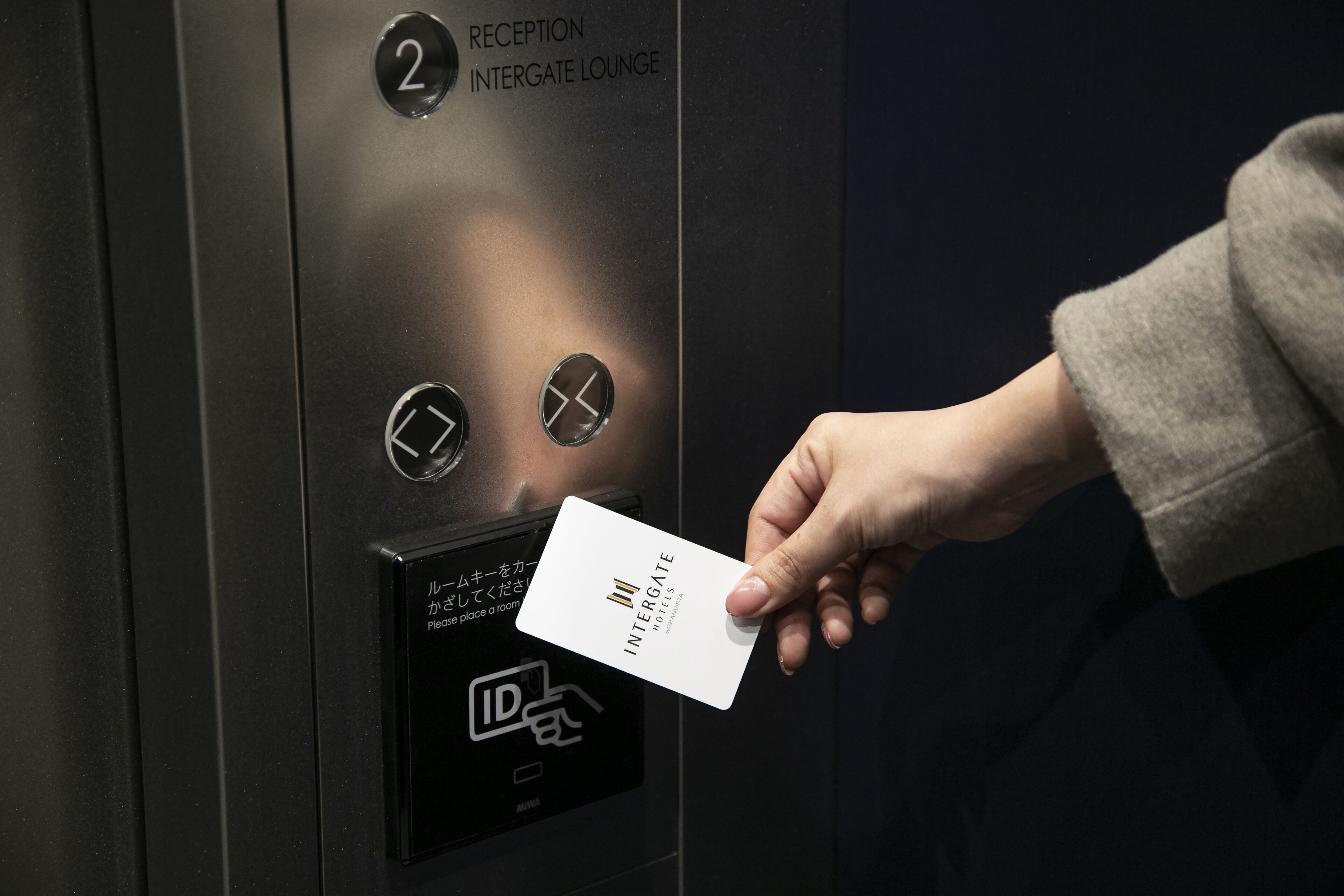 Una mano sosteniendo una tarjeta frente a un ascensor, con un letrero que indica 'Reception Intergate Lounge' y una ranura para identificaciones.