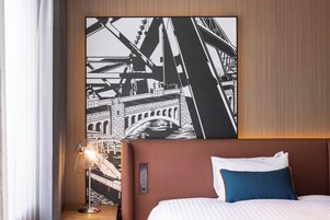 Ein Hotelzimmer mit einer großflächigen Schwarz-Weiß-Stadtlandschaftsmalerei, einem modernen Bett mit Kopfteil und einem Nachttisch mit Lampe.