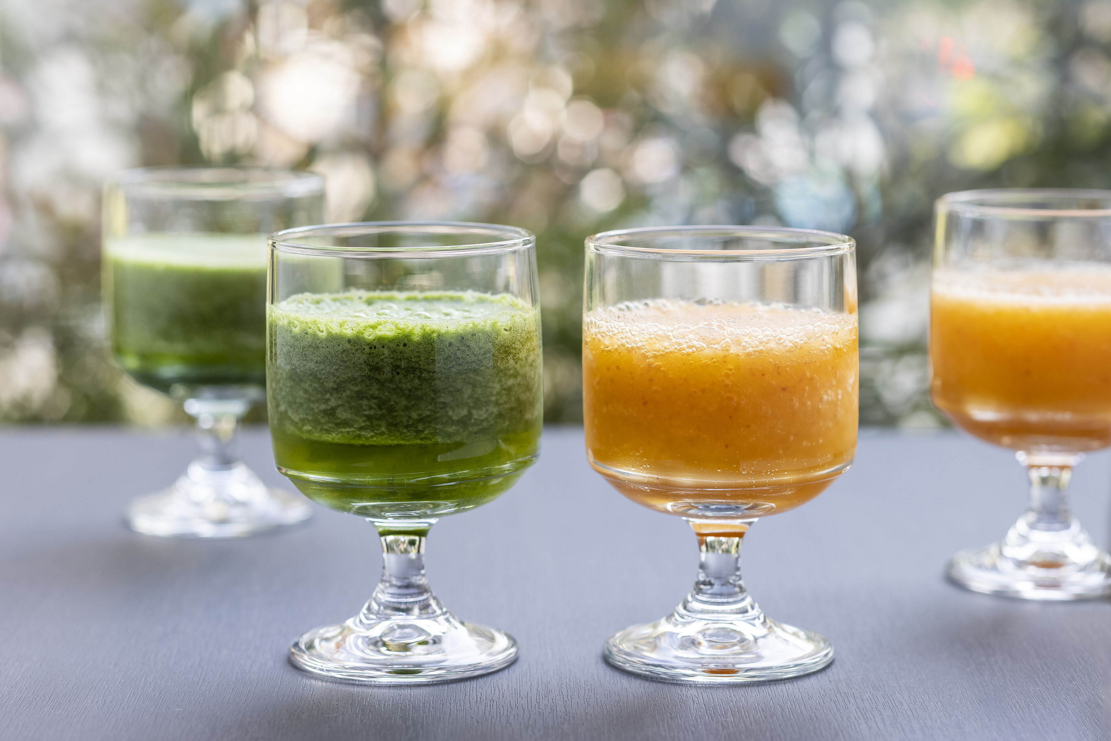 Tres vasos de bebidas verdes y anaranjadas sobre una mesa, con un fondo borroso al aire libre.