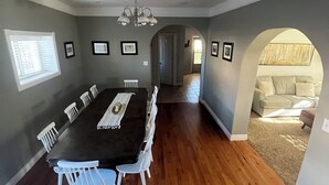 Un vaste espace dédié à la salle à manger, avec une longue table et des chaises blanches, un lustre, et une vue sur le salon à travers une arche.