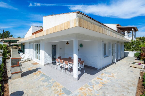 A covered patio with a table and chairs, a stone floor, and a white building with a tiled roof.