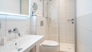 A modern bathroom with a white sink, mirror, and toilet. It includes a glass shower enclosure and a trash can. - Double room economy - lodging house oystercatcher (Norderney)