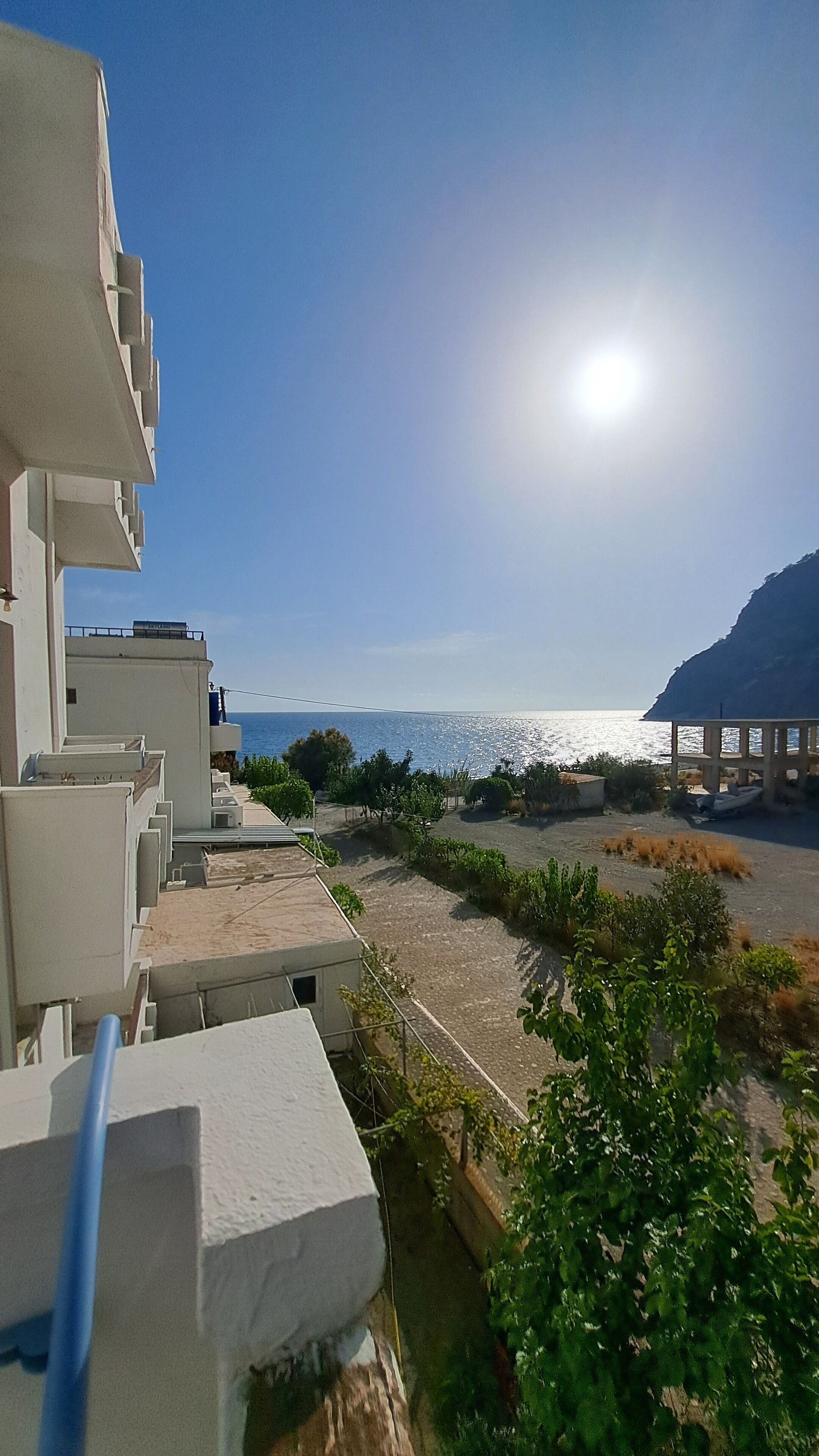 A coastal view with a clear sky, a sunlit sea, and a building with balconies overlooking the beach.