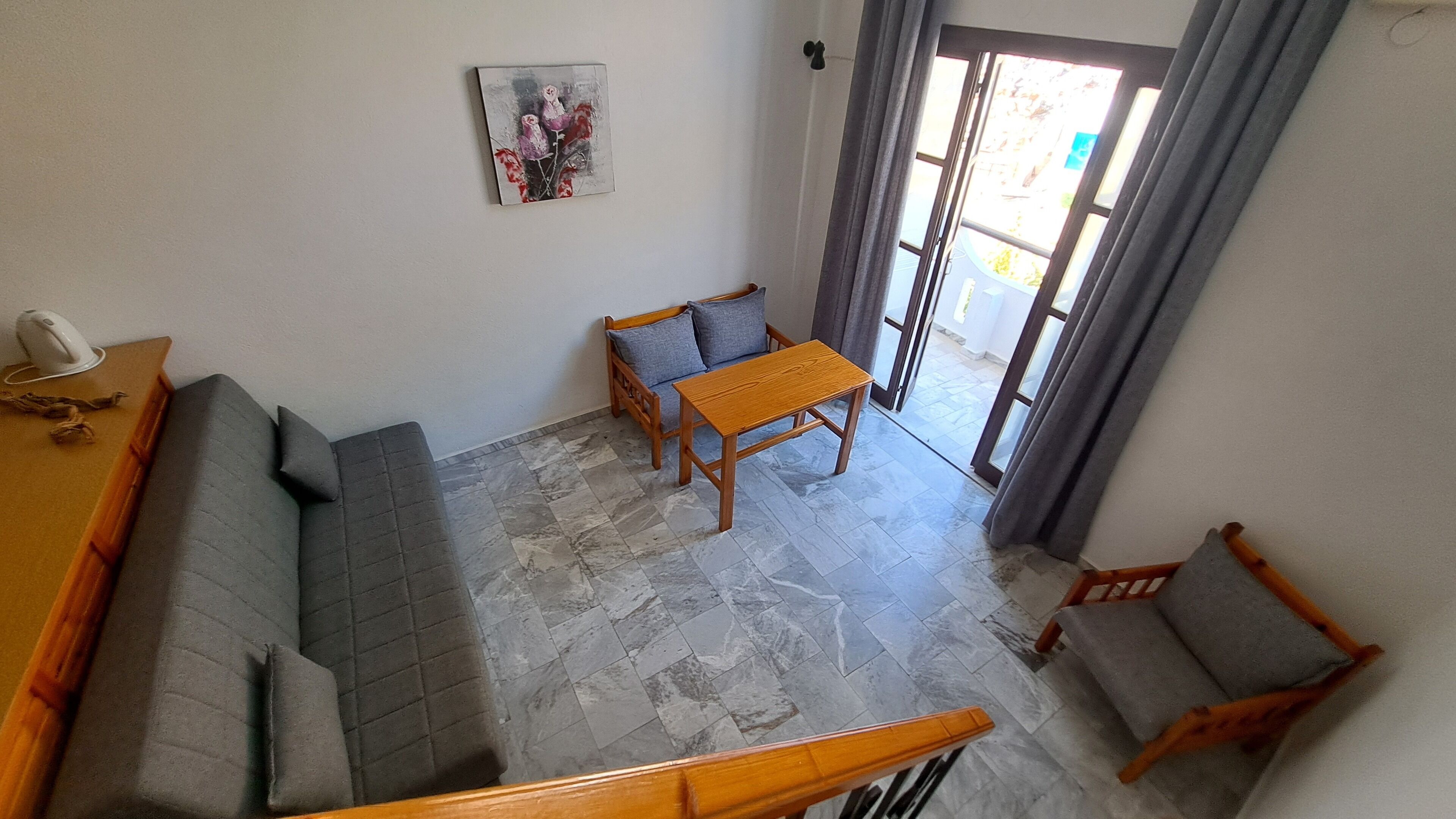 A living room with a sofa, a wooden table, and a chair. There is a painting on the wall and a door leading outside.