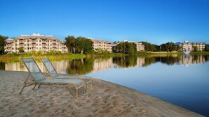 A serene lakeside scene with a sandy beach, two lounge chairs, and a row of multi-story buildings in the background. - 2 BR at Marriott Cypress Harbour (Orlando)