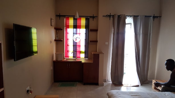 A bedroom with a bed, a TV mounted on the wall, a window with curtains, and a small kitchenette area. - Tranquil Corner Kampala (Kampala)