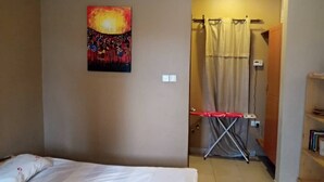 A hotel room with a bed, a painting on the wall, an ironing board, and a shower area. - Tranquil Corner Kampala (Kampala)