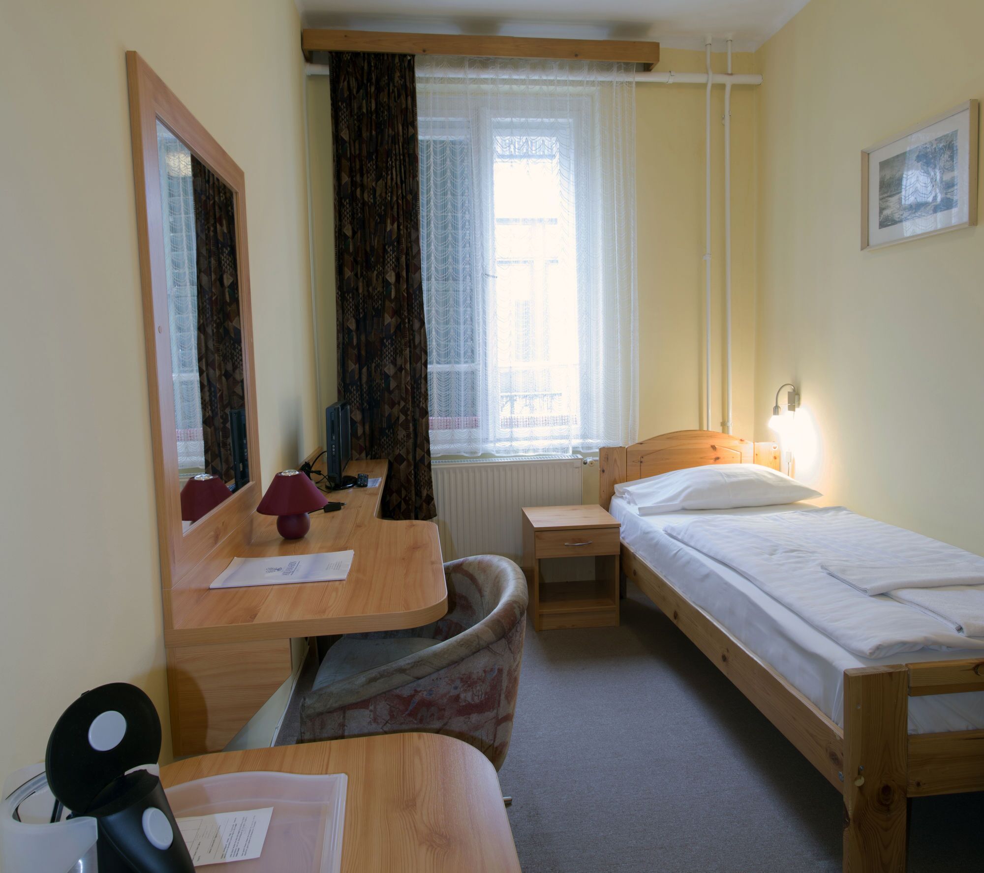 Una habitación de hotel pequeña y ordenada, con una cama individual, un escritorio con computadora, una silla, una mesita y una ventana con cortinas.