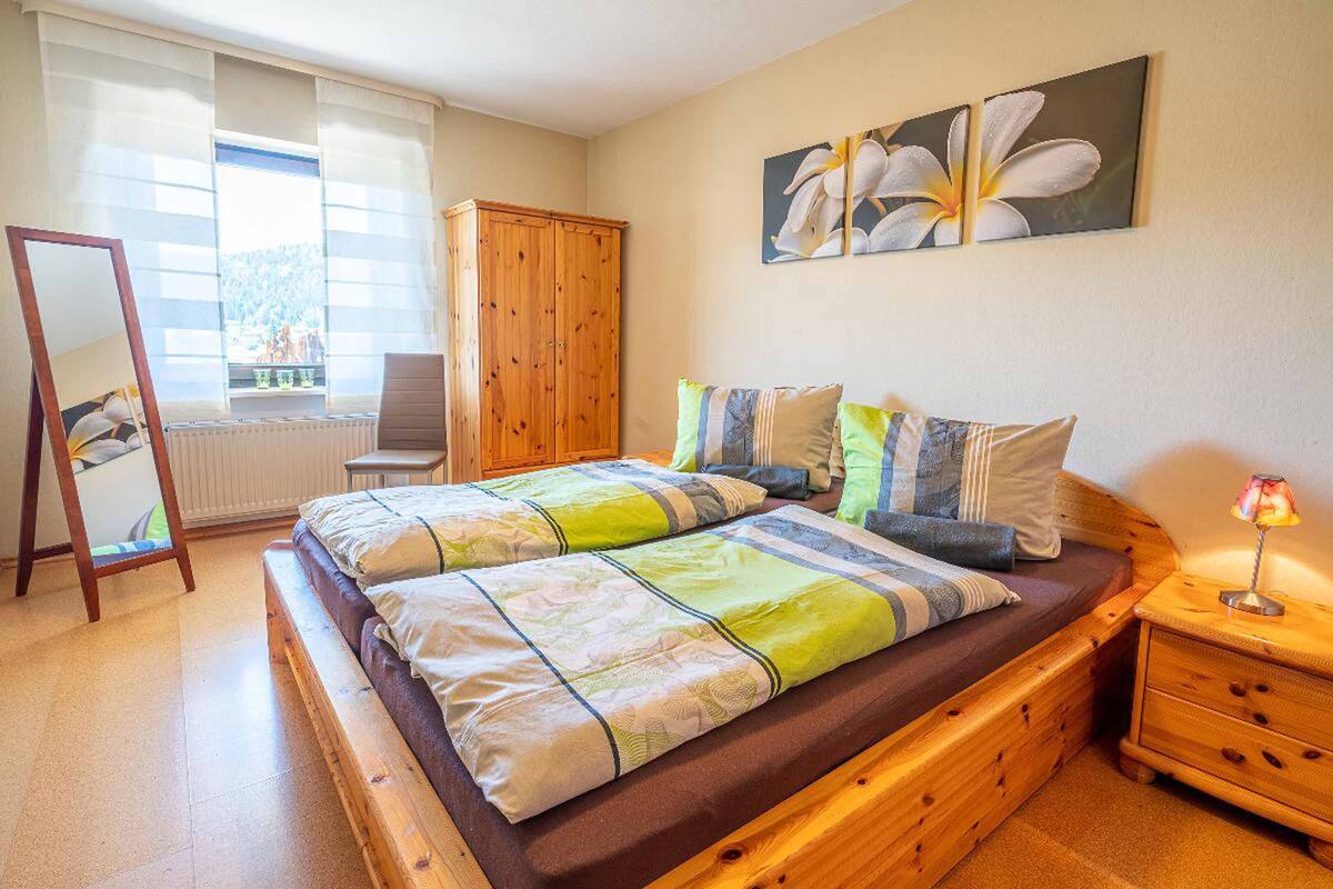 A bedroom with a wooden bed, a wooden wardrobe, a mirror, and a nightstand with a lamp.