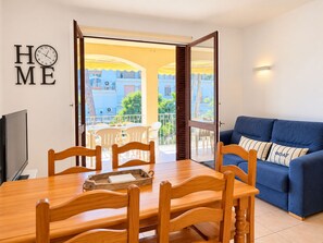 Ein Wohnzimmer mit blauem Sofa, Esstisch aus Holz und Stühlen, einem Fernseher sowie einem Balkon mit Blick auf Gebäude und Grünflächen.