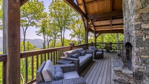 A wooden deck with wicker furniture, a stone fireplace, and a view of trees and mountains.