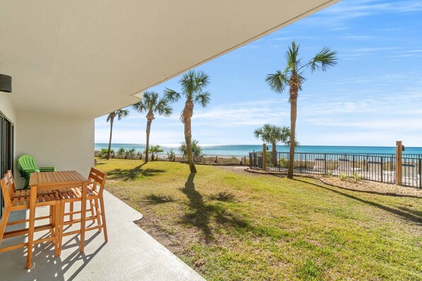 A balcony with a wooden table and chairs, overlooking a beach with palm trees and a clear sky.