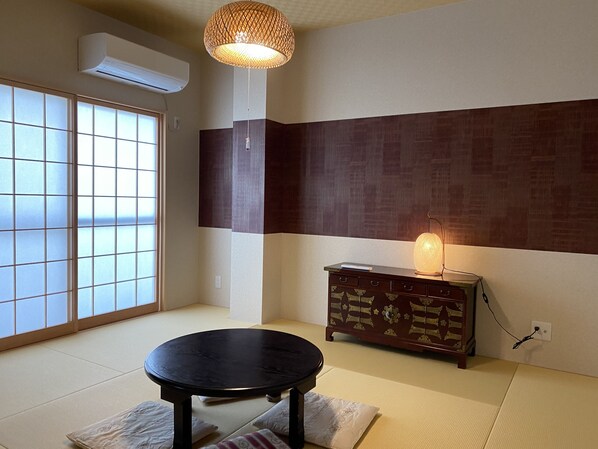 A traditional Japanese room with tatami flooring, a shoji screen, a tatami mat table, a wooden cabinet with a lamp, and a hanging lamp.