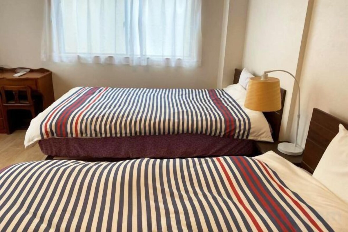 Habitación de hotel con dos camas, una mesita de noche, una lámpara y una ventana con cortinas translúcidas.