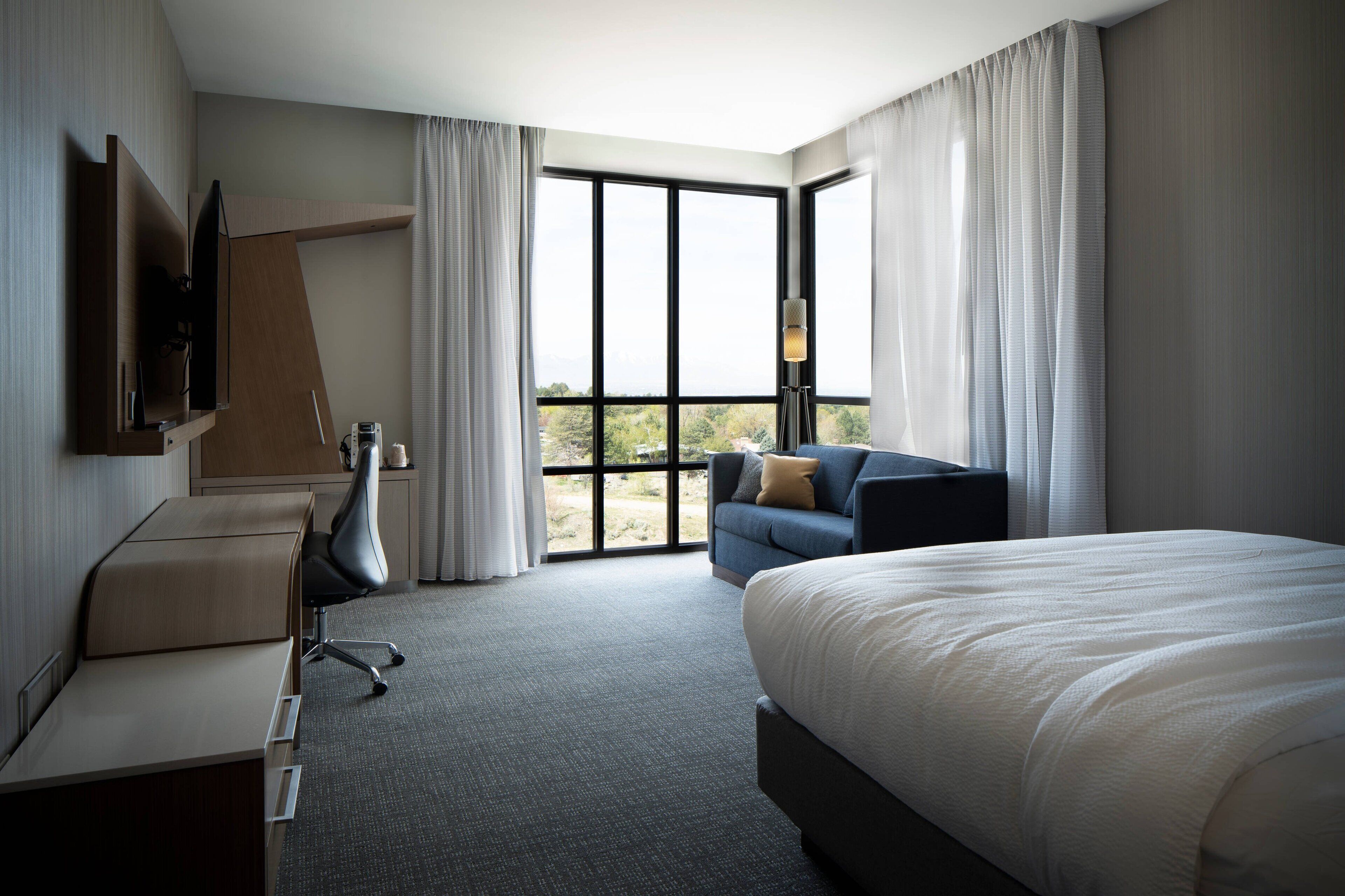A hotel room with a large bed, a desk, a chair, a sofa, and a TV. There are large windows with curtains.