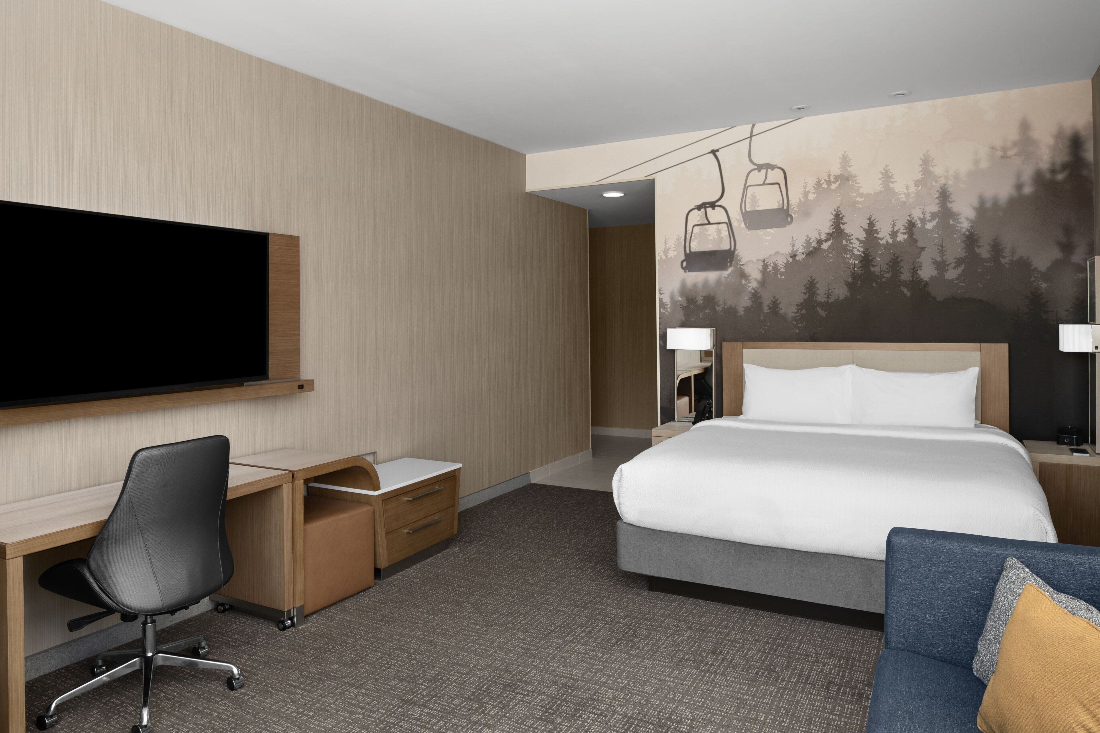 A hotel room with a bed, a desk with a chair, a TV, and a mural of a forest with a ski lift.